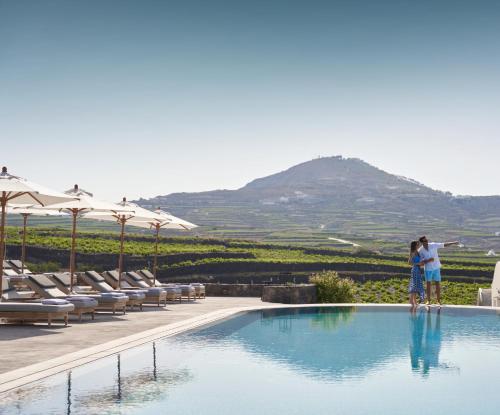 two people standing next to a swimming pool with a mountain at Vedema, a Luxury Collection Resort, Santorini in Megalokhori