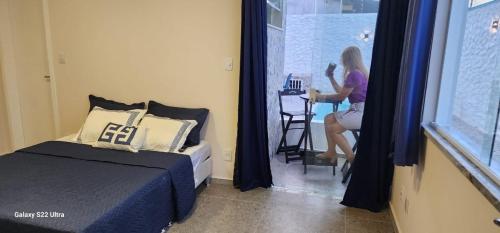 a woman sitting at a table in a room with a bed at Atena Flats in Aracaju
