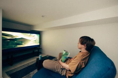mężczyzna siedzący na kanapie oglądający telewizję w obiekcie YOUR ROOM Kumamoto Station Little w mieście Hanabatachō