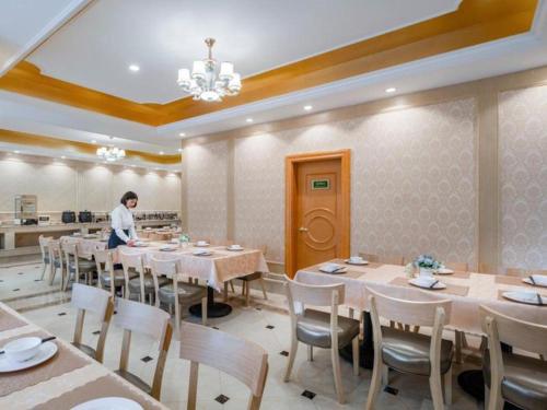 Vienna Hotel Kunming Dianzhong New District في Yanglin: رجل يقف في غرفة الطعام مع الطاولات والكراسي