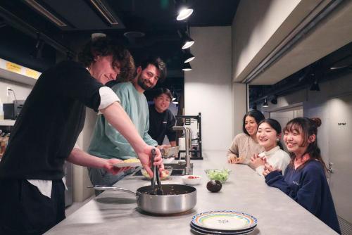 &AND HOSTEL SHINSAIBASHI EAST في أوساكا: رجل يعد الطعام في مطبخ مع مجموعة من الناس