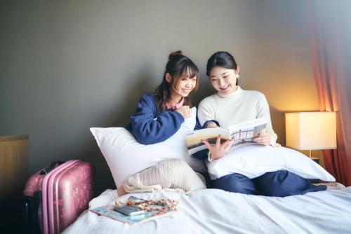 &AND HOSTEL SHINSAIBASHI EAST في أوساكا: كانتا جالستين على سرير يقرآن كتاب