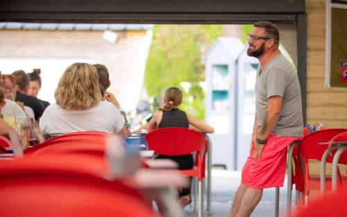 un hombre con pantalones cortos rojos parado frente a una mesa en Camping Colleville-sur-Mer, en Colleville-sur-Mer