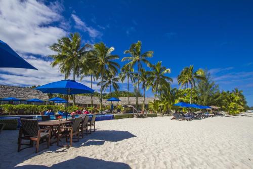 a beach with tables and chairs and blue umbrellas at Manuia Beach Resort in Rarotonga