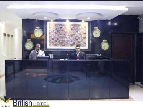 dos hombres sentados en un escritorio en una habitación con relojes en British Hotel - Johar Town LHR en Lahore