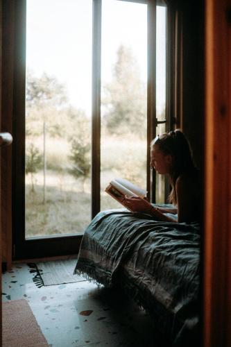 a woman sitting on a bed reading a book by a window at Maciejówka Borki 33 in Borki