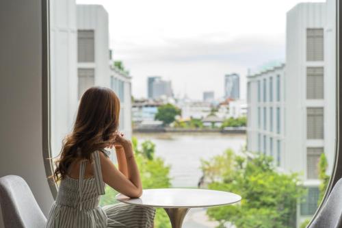 Una donna seduta a un tavolo che guarda fuori da una finestra di The Salil Hotel Riverside Bangkok a Bangkok