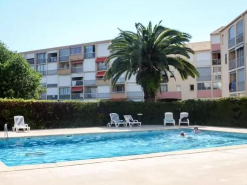 une piscine avec des chaises et un palmier ainsi qu'un bâtiment dans l'établissement Appartement avec accès piscine, proximité mer, au Grau-du-Roi
