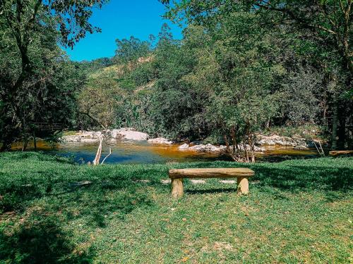 a wooden bench sitting in the grass near a river at Suíte com cachoeira no quintal in Antônio Carlos