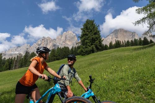 two people riding bikes on a hill with mountains in the background at Moseralm Dolomiti Spa Resort in Nova Levante