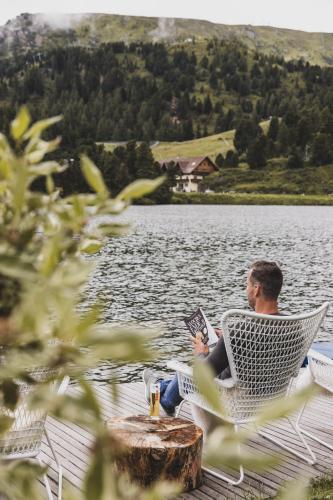 a man sitting in a chair reading a book by a lake at Seehotel Jägerwirt in Turracher Hohe