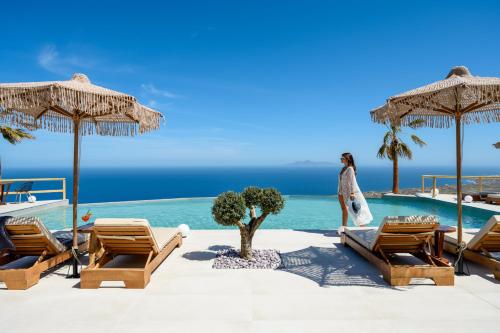 a woman standing next to a pool with chairs and umbrellas at Leon Luxury Suites in Imerovigli