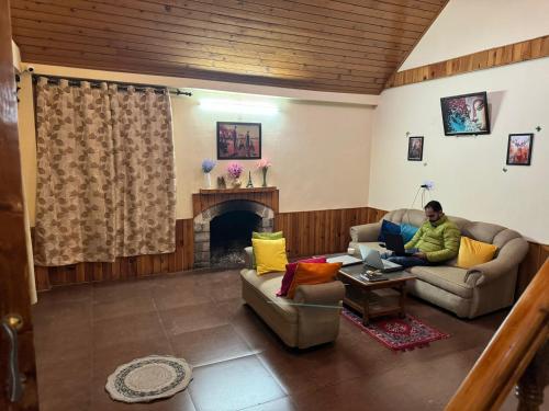 a man sitting on a couch in a living room with a laptop at Vamika Cottages in Manāli