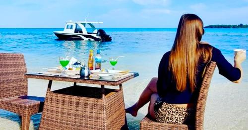a woman sitting at a table on the beach with a boat at Wind Breeze Sunset View Inn in Guraidhoo