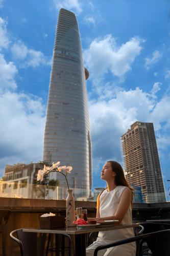 a woman sitting at a table in front of a tall building at Hato Saigon Boutique Hotel & Rooftop in Ho Chi Minh City