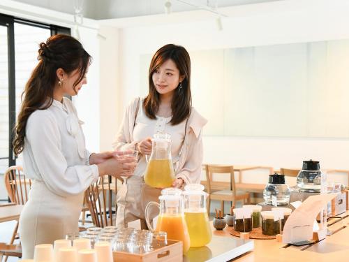 two women standing in a room with containers of orange juice at REF Kyoto Hachijoguchi by VESSEL HOTELS in Kyoto