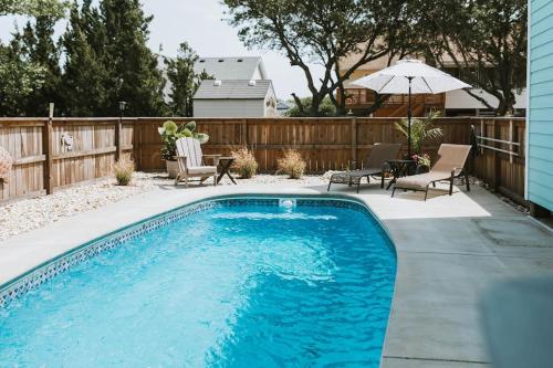 a swimming pool in a backyard with chairs and an umbrella at Somewhere On A Beach - Dog Friendly • Private Pool • Hot Tub • Fire Pit • Game Room • Horseshoe Pits • Walk to beach, Sound, arcade and go carts! in Kill Devil Hills