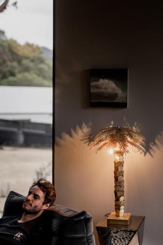 a man sitting in a chair next to a table with a lamp at 'Sisters' - Cliff top cabin with hot tub in Spring Beach