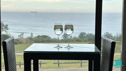two wine glasses on a table with a view of the ocean at Ripples by the Sea in Caves Beach