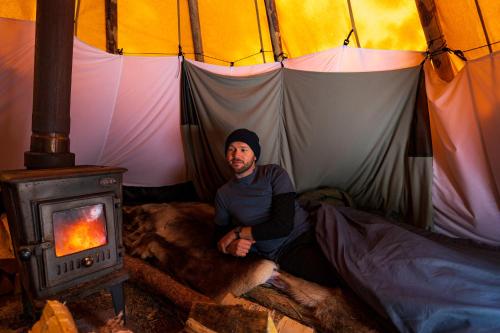 a man sitting in a tent with a wood stove at Reindeer Lodge in Jukkasjärvi