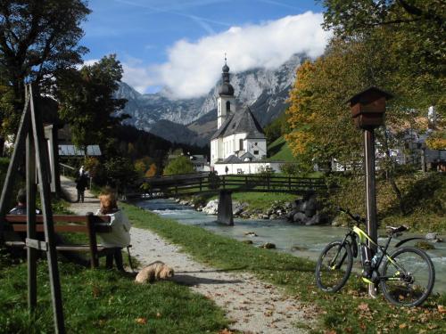 a woman sitting on a bench next to a river with a church at Haus Buchenwald in Ramsau