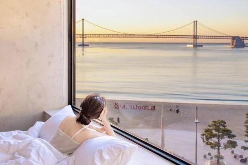 a woman sitting on a bed looking out a window at a bridge at Paradise on earth in Busan