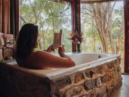a woman sitting in a bath tub reading a book at Firefly Falls in Plettenberg Bay