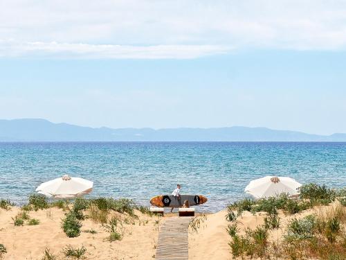 a man sitting on a beach with a surfboard and two umbrellas at Mandola Rosa at Riviera Olympia, A Grecotel Resort to Live in Loutra Killinis