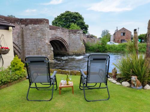two chairs sitting in the grass next to a bridge at River Cottage in Penrith