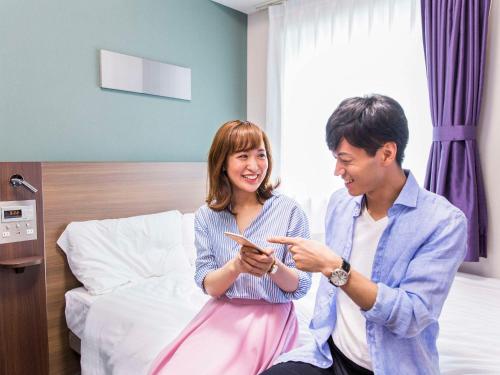 a man and a woman sitting on a bed looking at a cell phone at Comfort Hotel Obihiro in Obihiro