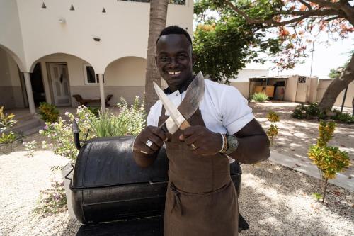 a man in a brown apron holding two knives at La Teranga Apartament Parrot in Toubakouta