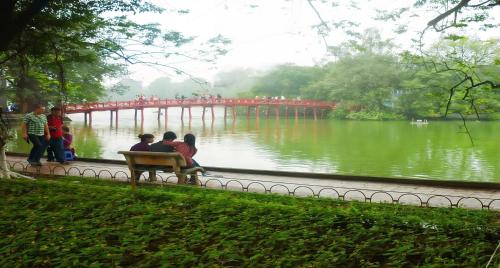 three people sitting on a bench near a river with a bridge at Hanoi Win Hotel & Travel in Hanoi
