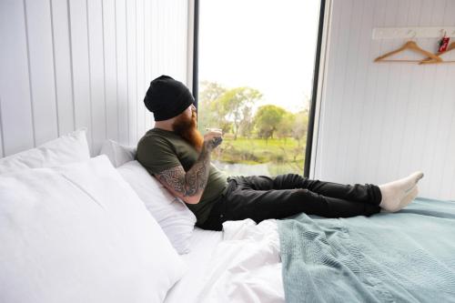 a man sitting on a bed looking out a window at McCabe Farm Cabin by Tiny Away in Brookton