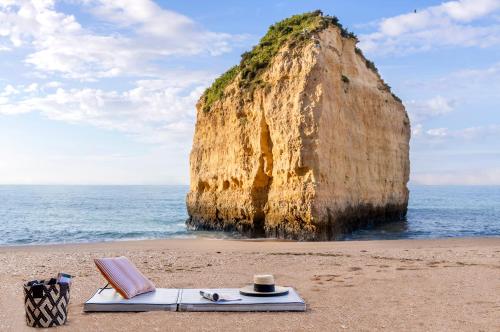 a hat and a book on a beach with a large rock at Vilalara Grand Hotel Algarve in Armação de Pêra