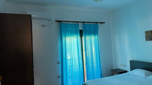 a bedroom with a bed and a window with blue curtains at Amel's Apartments Comfortable Family Stay Near Orikum Beach in Orikum