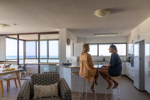 two women are sitting in a kitchen with a counter at Breezy Kings Beach Apartment with Ocean Views in Caloundra