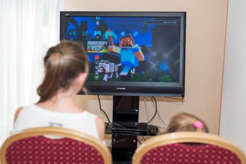 a woman and a child sitting in front of a television at Hotel Kudowa Manufaktura Relaksu in Kudowa-Zdrój