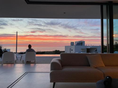a person sitting at a table in a living room with a sunset at Exclusive Beach House with full sea view, Luxury and Comfort by the Sea in Murtinheira