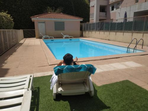 un homme assis dans une chaise à côté d'une piscine dans l'établissement Studio avec coin nuit et garage, à Saint-Laurent-du-Var