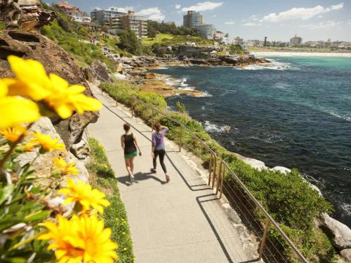 two people walking down a sidewalk next to the ocean at Beachside bright unit - sleeps 4! in Sydney