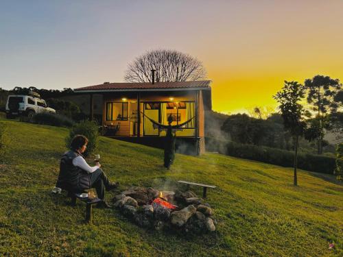 a man sitting in front of a fire in front of a cabin at 4 Estações & Chalé da Pedra in Sapucaí-Mirim