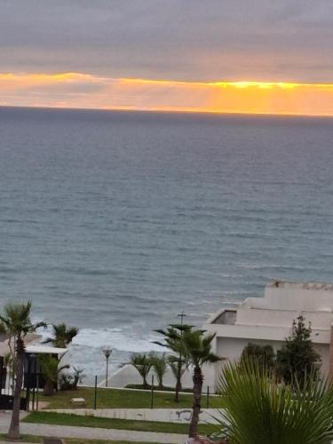 a sunset over the ocean with palm trees and a building at Appt magnifique vue mer et piscine spacieuse in Sidi Bouqnadel