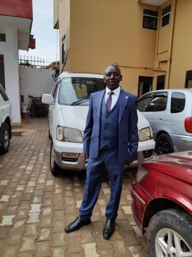 Un hombre con traje y corbata de pie delante de los coches. en BK EXECUTIVE HOTEL Nansana, en Wakiso
