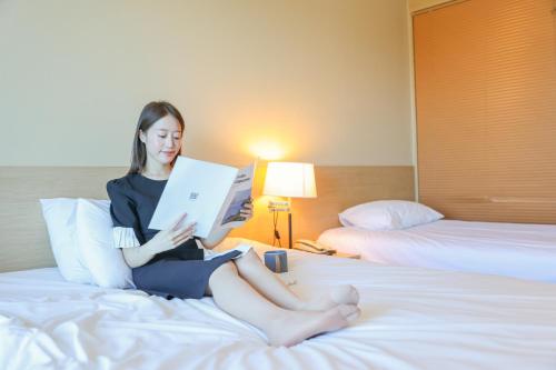 a woman sitting on a bed reading a book at From H The Best Hotel Jeju Seongsan in Seogwipo