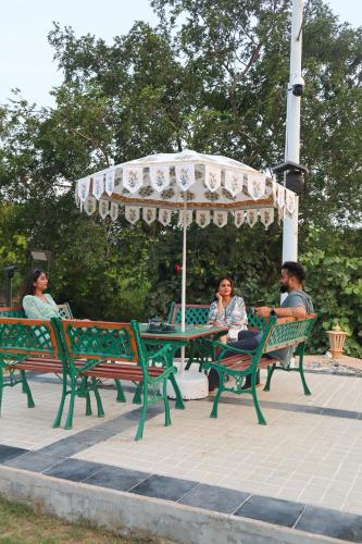 three people sitting at a picnic table under an umbrella at Elivaas 5 BHK with Pool, Tents & Pond - Kaimalgarh Farmstay in Bhundsi
