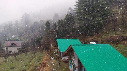 a snow covered hillside with a green building and trees at The Satvic Retreat in Manāli
