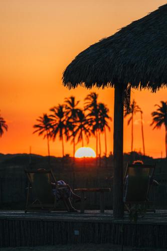 two chairs and an umbrella and palm trees at sunset at Vila Ronquinha - Arpoador in Tutóia
