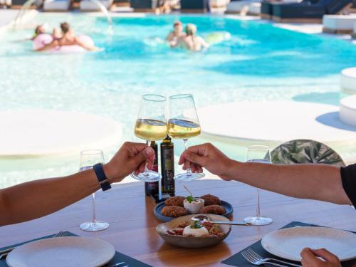 two people holding wine glasses at a table near a swimming pool at Axel Beach Mykonos - Adults Only in Mýkonos City