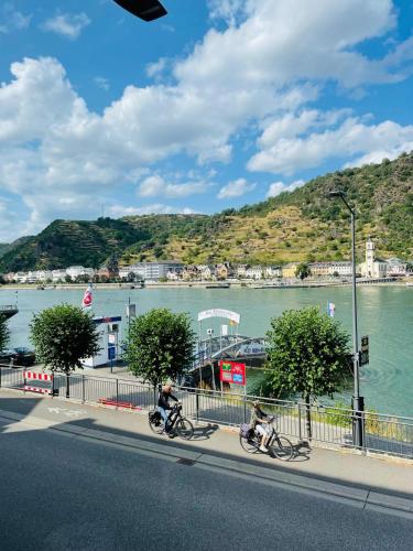 two people riding bikes down a street next to a river at Rheinromantik mit Blick auf die Burg Katz, bis 4P in Sankt Goar