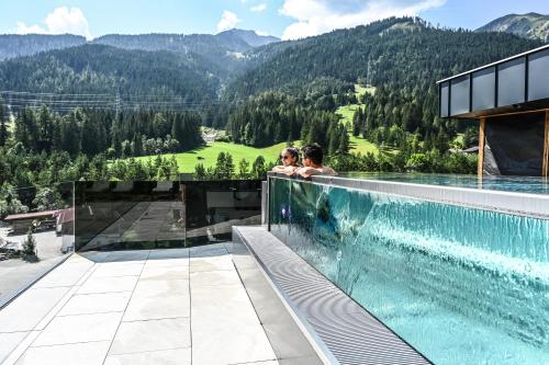 two people sitting on the edge of a swimming pool overlooking a mountain at Hotel Lärchenhof in Lermoos